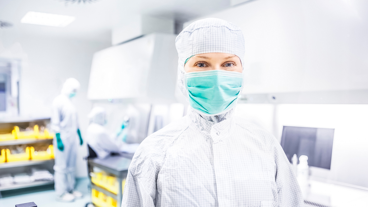 Eine Mitarbeiterin mit Schutzanzug und Maske steht im Reinraumlabor und blickt in die Kamera. Im Hintergrund arbeiten zwei Kollegen an einer Werkbank und stellen eine Infusion her.